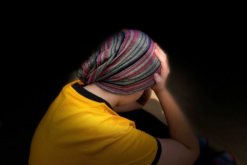 Woman in yellow shirt sits with head, tied in headscarf, in her hands, with black background.