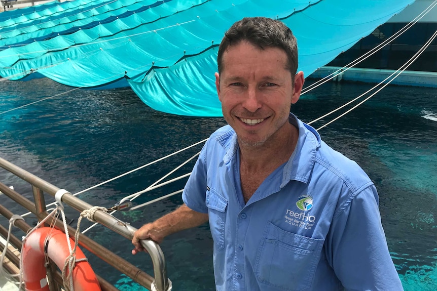 Smiling man wears ReefHQ shirt and holds guard rail as he stands in front of large aquarium pool