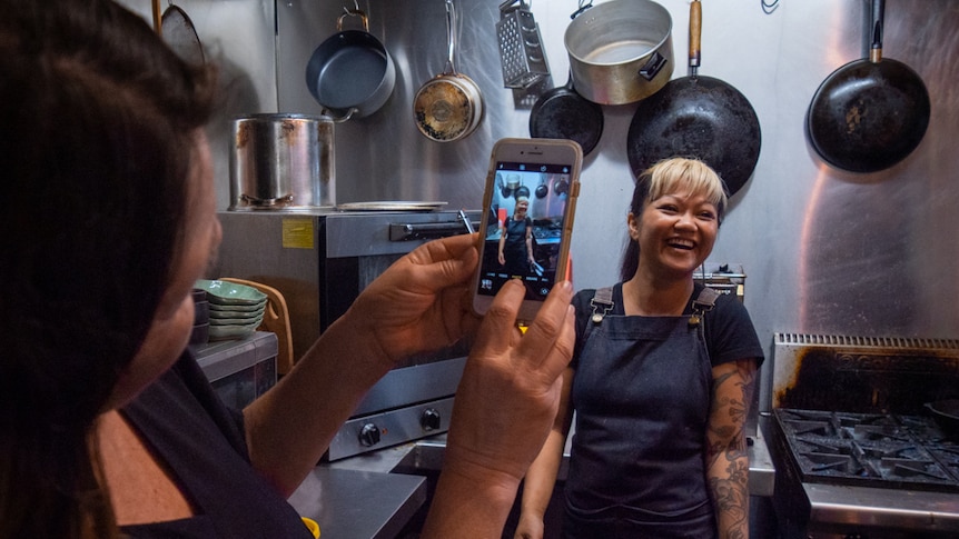 An artist takes a photo of cook Busara O'Reilly who stands in her cafe kitchen surrounded by pots and pans