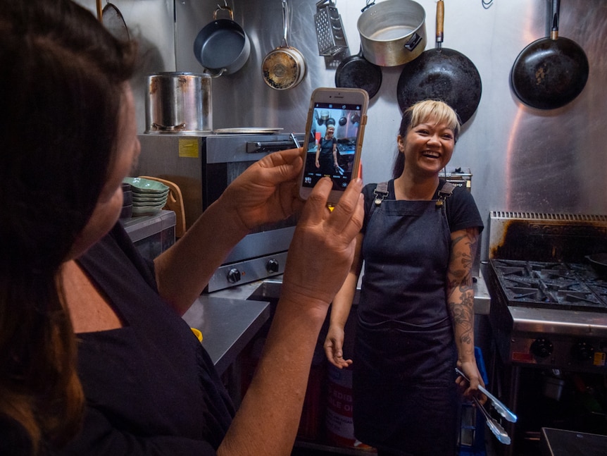 An artist takes a photo of cook Busara O'Reilly who stands in her cafe kitchen surrounded by pots and pans