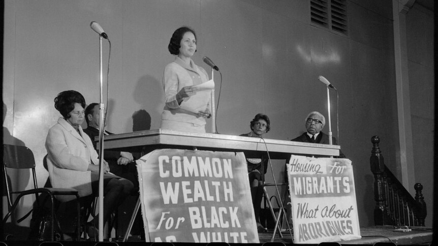 A black-and-white photo of a woman speaking at a microphone with protest signs beneath the table.