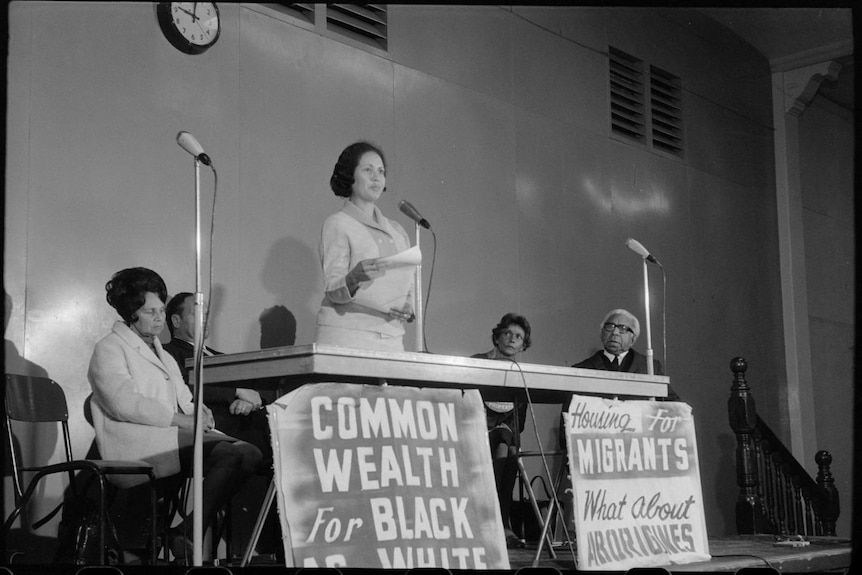 A black-and-white photo of a woman speaking at a microphone with protest signs beneath the table.