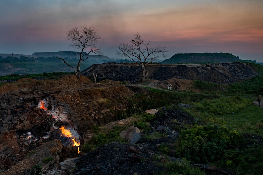 A view of burning coal in the foreground at dusk with trees and plain in the background.