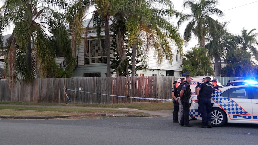 Police standing around a police car in front of a green house with palm trees and police tape