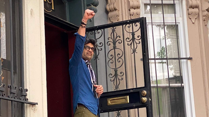 A man of Indian descent raises his fist in the doorway of an ornate, stone townhouse.