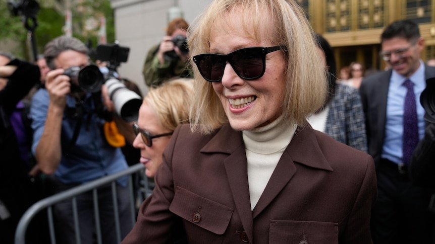 A woman with blonde bobbed hair, dark sunglasses and brown jacket smiles outside court