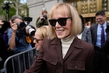 A woman with blonde bobbed hair, dark sunglasses and brown jacket smiles outside court