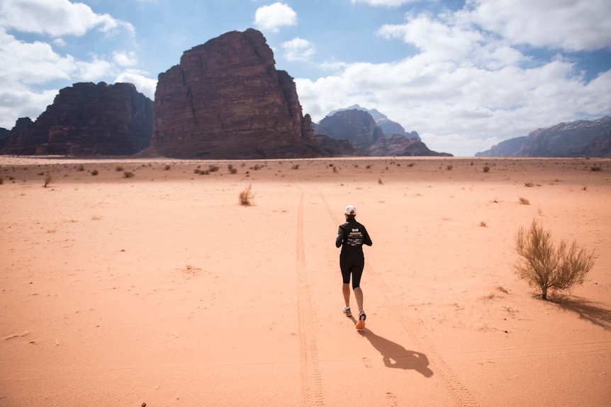 A woman runs across a sand desert where large rock formations can be seen in the background