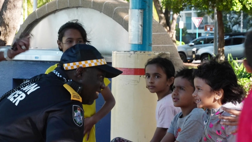 A man in a uniform talks to a group of kids, smiling
