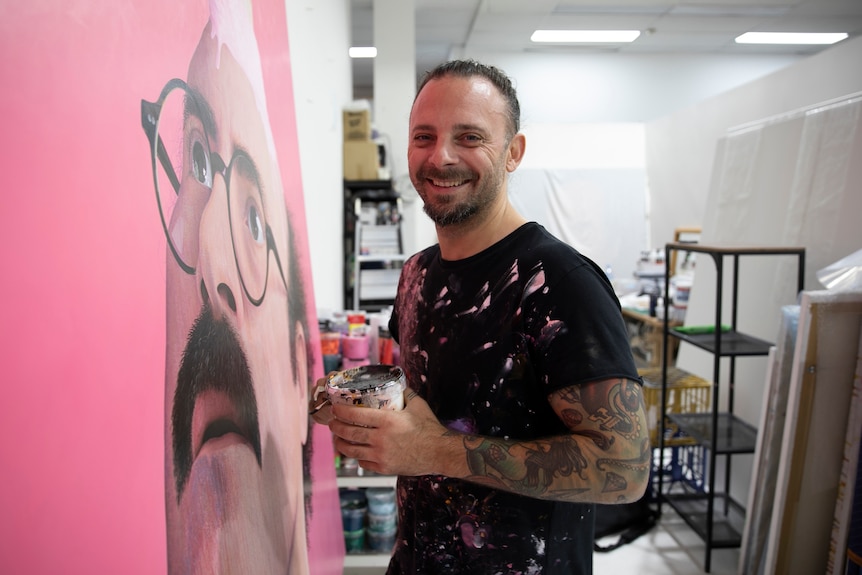 a smiling man holding a round object looking standing in front of a large picture of a man with hair on his face wearing glasses