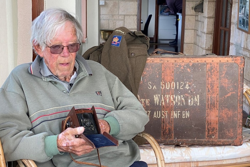 Old man looking out of shot holding camera a box brownie camera, army jacket and suitcase in background