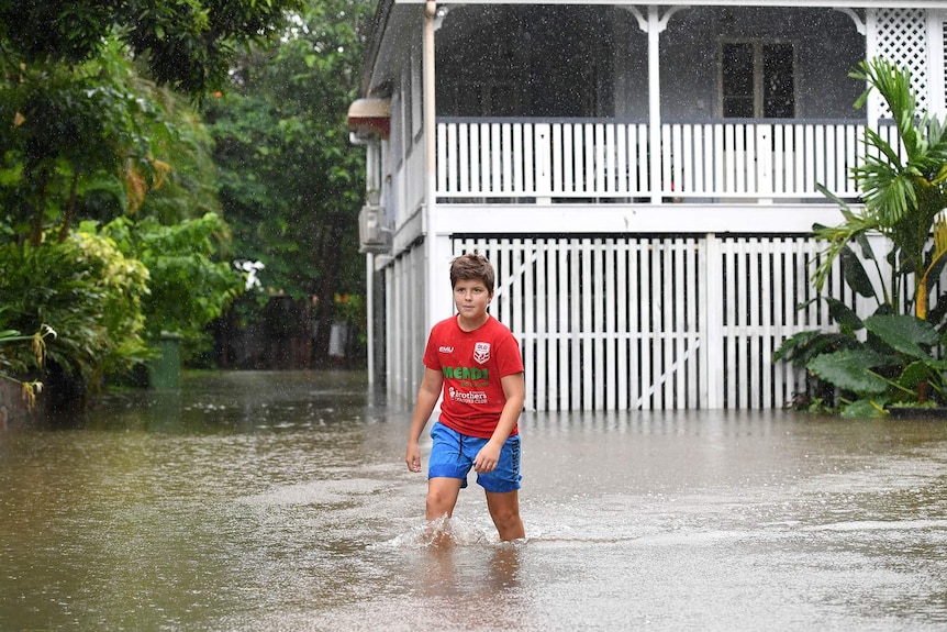 A boy wading through floodwater outside an old Queenslander house