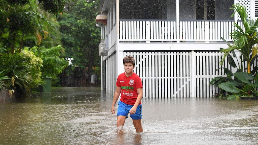 A boy wading through floodwater outside an old Queenslander house