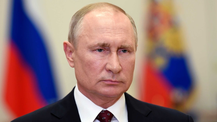 A man in a suit looks ahead as he stands in front of two flags.