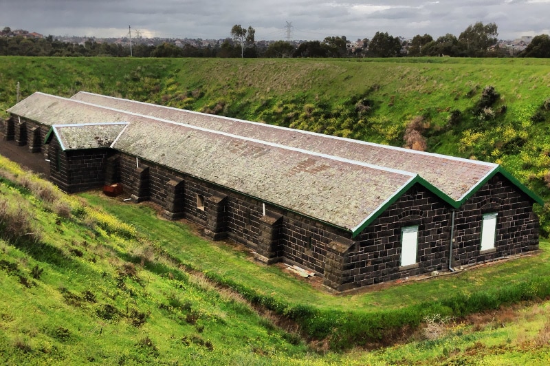 A long, rectangular, bluestone building with an "M" shaped roof, surrounded by tall, grassy mounds.
