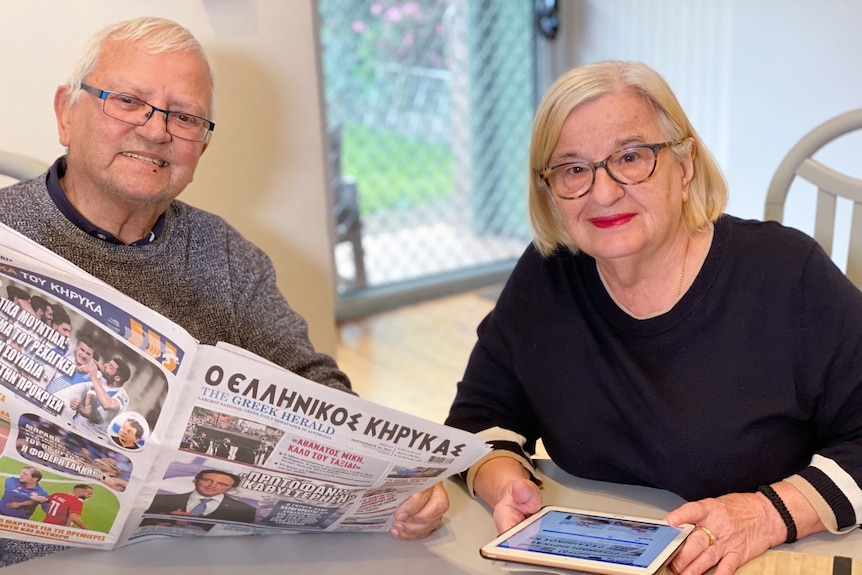 A smiling man reading a Greek newspaper next to a woman reading from an iPad.