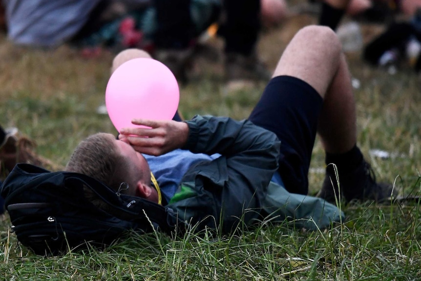 A festival-goer lying on the ground on his back inhaling nitrous oxide through a balloon