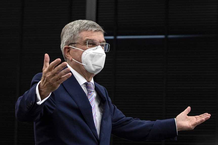 A mask-wearing sports executive in a suit gestures as he makes a speech.