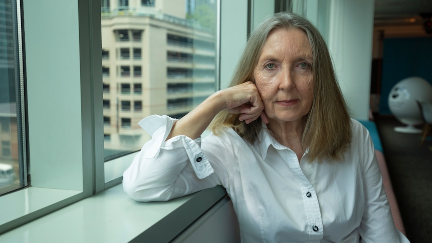 Miranda Jakich, with stern expression, white shirt and shoulder-length fair hair, leans a hand to her face, sitting near window.