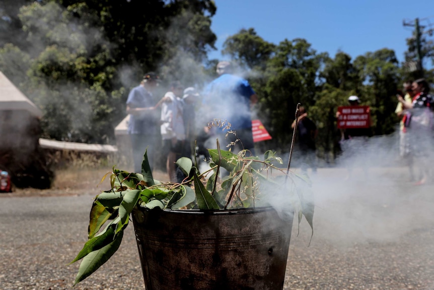 A small tin bucket with smoking eucalypt leaves sits on large driveway with people holding signs visible in background