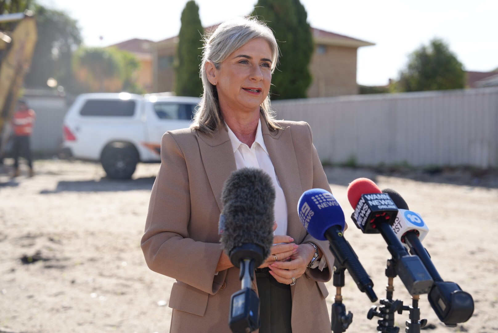 Sandra hablando en una conferencia de prensa al aire libre con una camisa blanca y una chaqueta de biegio  