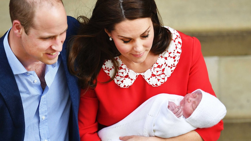 A man in a blue shirt and jacket has him arm around a woman in a red dress holding a baby wrapped in white fabric