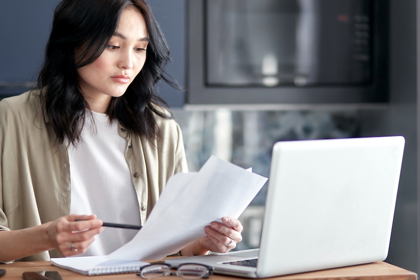 A woman with a confused look holds papers while looking at a laptop.