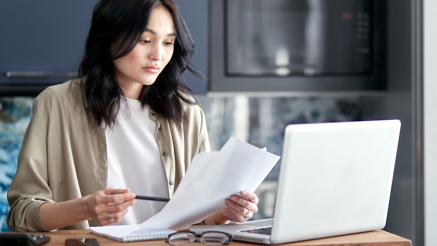 A woman with a confused look holds papers while looking at a laptop.