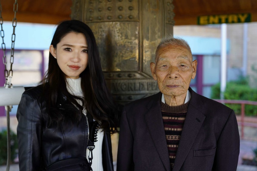 An elderly man and a young woman with long black hair stand in front of a large bell.