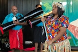 A woman wearing a colourful floral shirt dances while a man plays a ceremonial drum in the background.