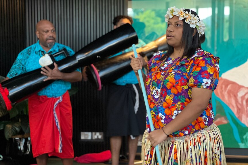 A woman wearing a colourful floral shirt dances while a man plays a ceremonial drum in the background
