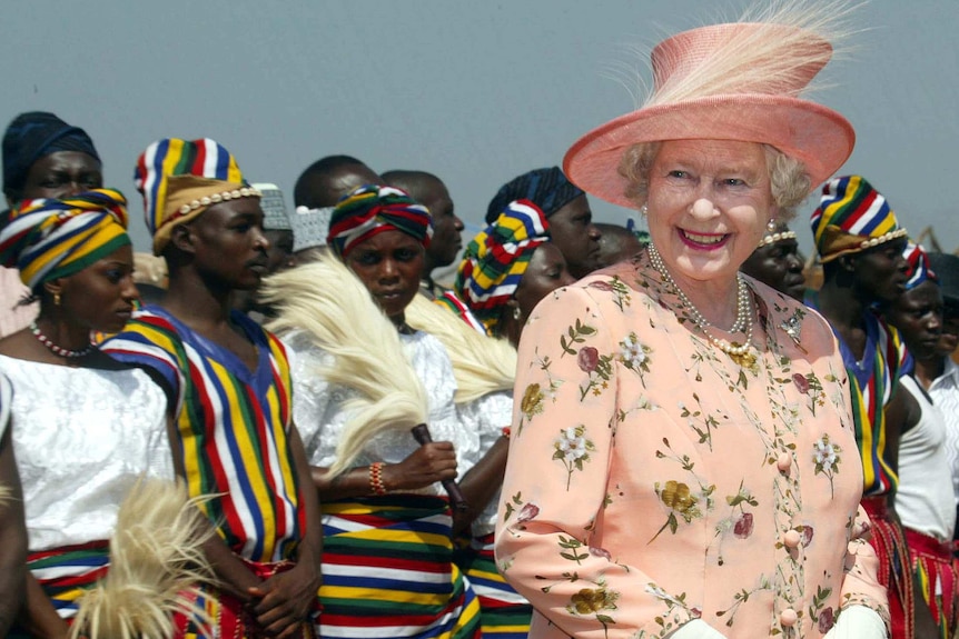 Queen Elizabeth II smiles as people in traditional dress stand behind her in Nigeria.