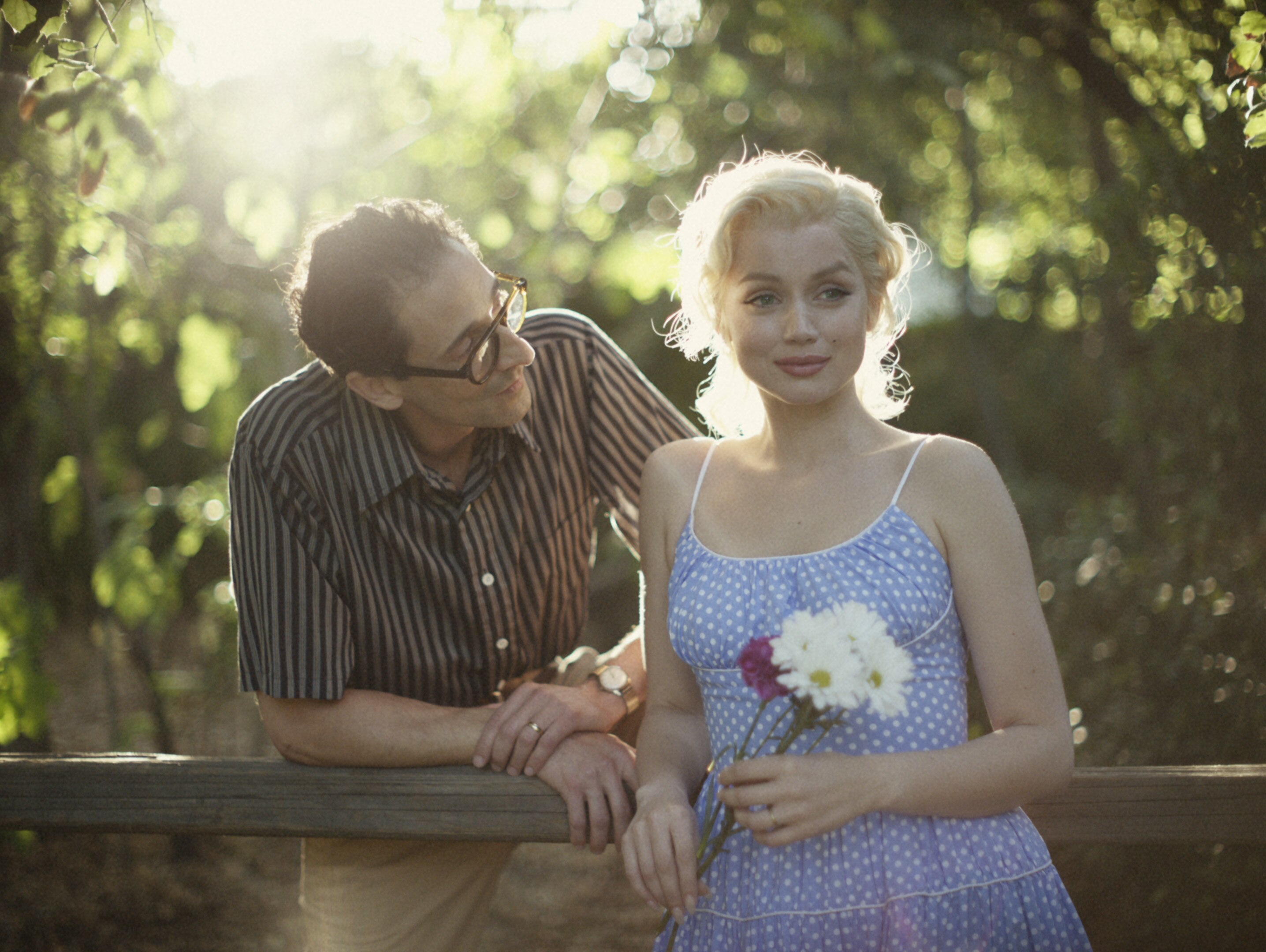 White man with dark hair, striped shirt and glasses leans on fence with white blonde-haired woman in blue dress holding flowers.
