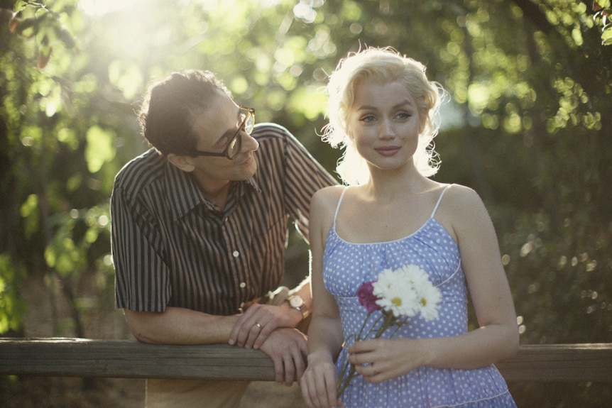 White man with dark hair, striped shirt and glasses leans on fence with white blonde-haired woman in blue dress holding flowers.