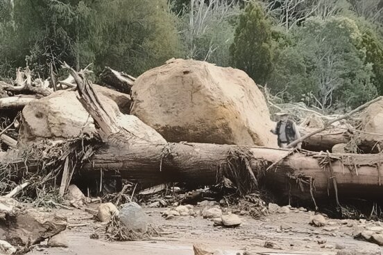 A man stands next to an enormous boulder among flood debris on a road