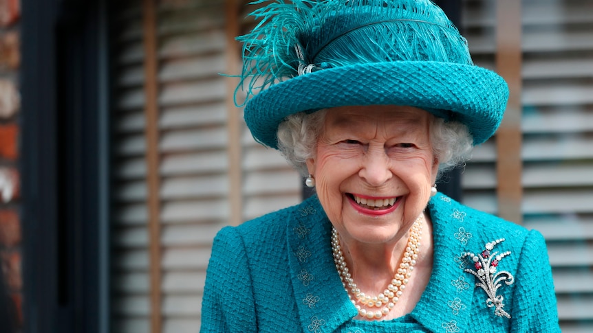 Queen Elizabeth II wearing a blue outfit and hat smiles toward the camera