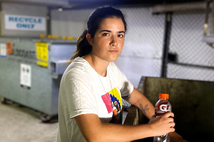 A woman with dark hair leans against a dumpster. 