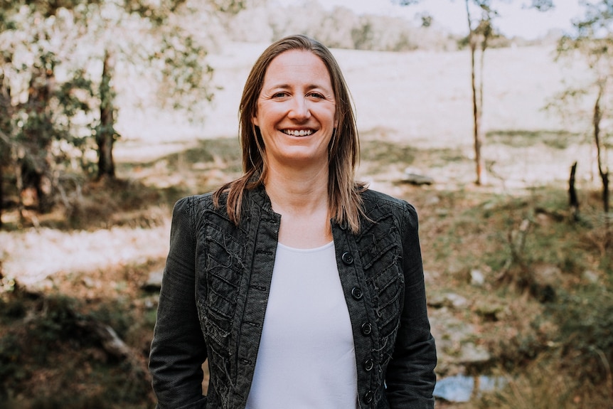 A woman in dark jacket stands smiling in a garden.