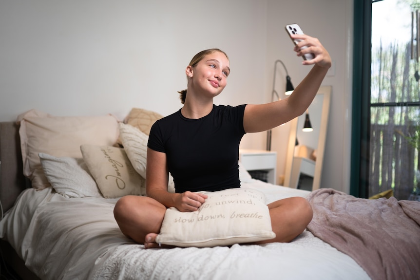 A teenage girl with blonde hair sitting cross-legged on a bed and taking a selfie.