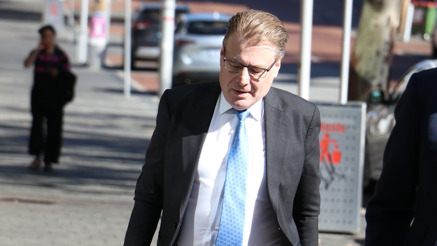 A mid-shot of WA Ombudsman Chris Field walking into a building wearing spectacles, a dark grey suit, blue tie and white shirt.
