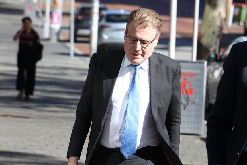 A mid-shot of WA Ombudsman Chris Field walking into a building wearing spectacles, a dark grey suit, blue tie and white shirt.