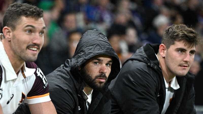 A dejected Brisbane Broncos player sits between teammates on the bench with the hood up on his jacket, looking at the game.