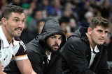 A dejected Brisbane Broncos player sits between teammates on the bench with the hood up on his jacket, looking at the game.