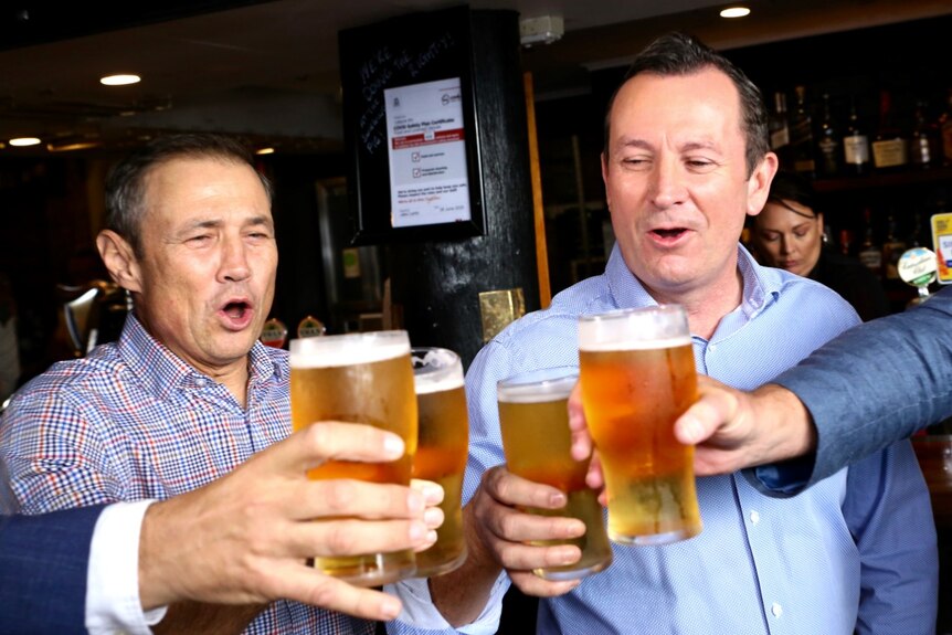 Two men holding pints of beer make a 'cheers' gesture with other people at a pub.