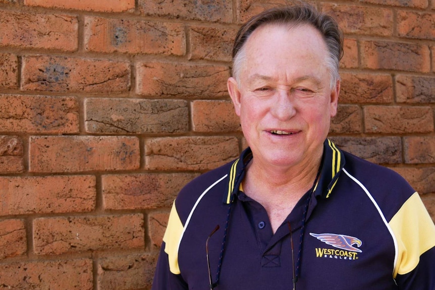An older Caucasian smiling man with greying hair,  in front of a brick wall, purple and cream shirt with logo.