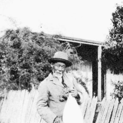 An old black and white photo of a bearded man in a hat and suit lifting up a bag. A picket fence and house are in the background