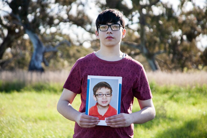 A teenager in glasses holds up a school photo of himself at six years old.