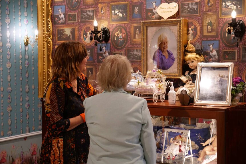 Two women smiling at a portrait hanging on the wall at a doll museum.