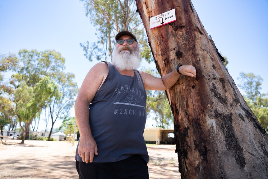 A man has a bushy beard, wears a dark coloured tank, hat and sunnies, he leans against tree with flood marker