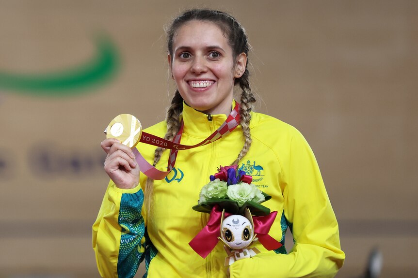 A woman in a green & gold tracksuit smiles holding up a gold medal that hangs from her neck. She holds flowers & an official toy